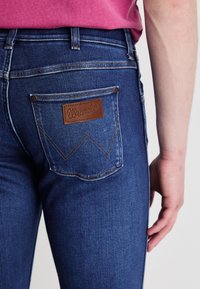 Donkerblauwe spijkerbroek van denim met een bruine leren Wrangler-patch en contrasterende stiksels. Beschikt over een klassiek vijf-pocket ontwerp en een slim fit.