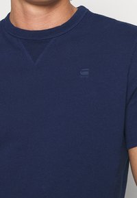 Camiseta de algodón azul marino con cuello redondo y mangas cortas. Presenta un logo sutil en la esquina superior izquierda y costuras planas.