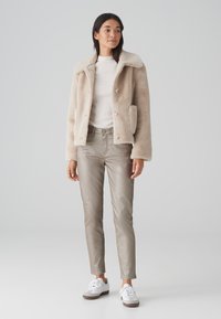 Veste en fausse fourrure beige avec un col, des boutons devant et des poches latérales. Associée à un pantalon beige métallique et des baskets blanches.