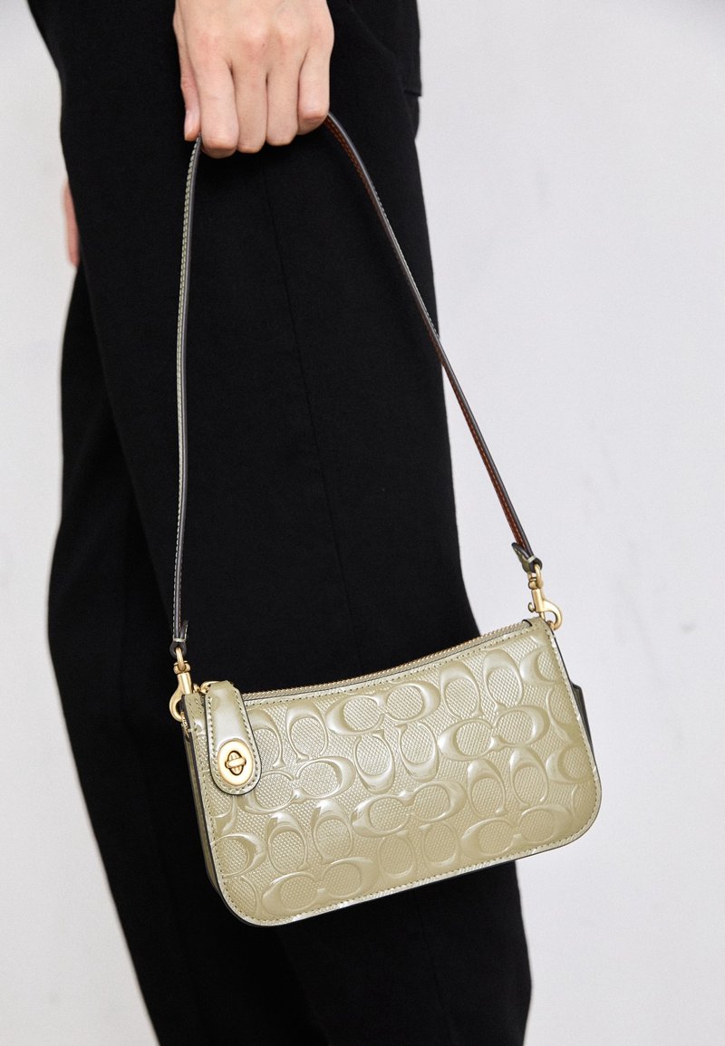 Main tenant un petit sac à main en cuir beige embossé avec une courte bandoulière foncée sur fond de pantalon noir.