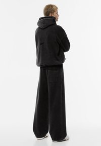 Veľká čierna mikina s textúrovaným povrchom a kapucňou, v kombinácii s voľnými čiernymi nohavicami s zadnou vreckom. Sú viditeľné úhľadné detaily šitia.