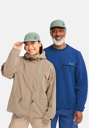 Smiling man and woman wearing outdoor jackets and light green caps, posing against a plain light background.