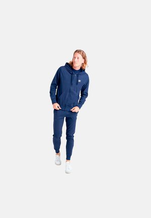 Homme aux cheveux mi-longs, portant un sweat à capuche zippé bleu marine et un pantalon de jogging, marchant vers l'avant les mains près des poches et des baskets blanches.