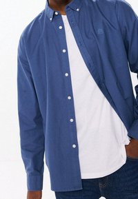 Koszula na guziki w ciemnoniebieskiej bawełnie, z długimi rękawami, o luźnym kroju, z białymi guzikami i małym logo na piersi, noszona na białej koszulce.
