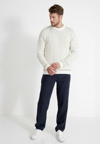 Witte gebreide trui met een patroonstructuur, ronde hals, gecombineerd met donkerblauwe broeken en witte sneakers. Eenvoudige, casual outfit.