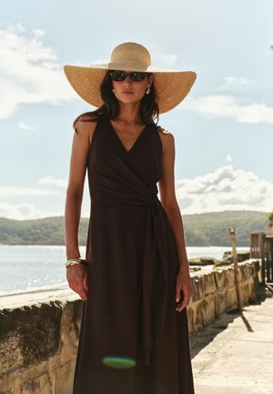 Mujer con un sombrero de paja de ala ancha, vestido oscuro sin mangas, gafas de sol y joyas de oro, de pie junto a una barandilla de piedra cerca del agua en un día soleado.