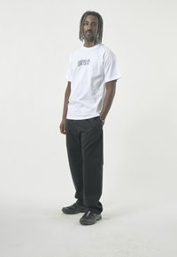 White t-shirt featuring a colorful graphic design, paired with black pants and black athletic shoes, set against a neutral background.