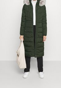 Zielona kurtka puchowa z szarą kapturem wyłożonym futrem, pionowym przeszyciem, suwakiem z przodu i kieszeniami po bokach. Noszona z białą bluzką i czarnymi spodniami.