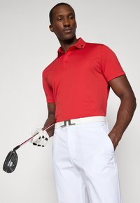 Camisa polo vermelha de mangas curtas e colarinho, combinada com calças brancas. O modelo segura um taco de golfe, vestindo um cinto e uma luva brancos.