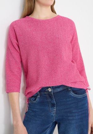 Mujer vistiendo un suéter tejido de textura brillante en color rosa intenso con mangas tres cuartos, combinado con jeans de mezclilla azul oscuro.