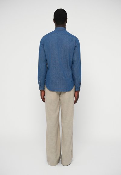 Világoskék farmer ing hosszú ujjakkal és gombos mandzsettával, laza szabású bézs nadrággal párosítva. Egyszerű kialakítás, látható minták nélkül.