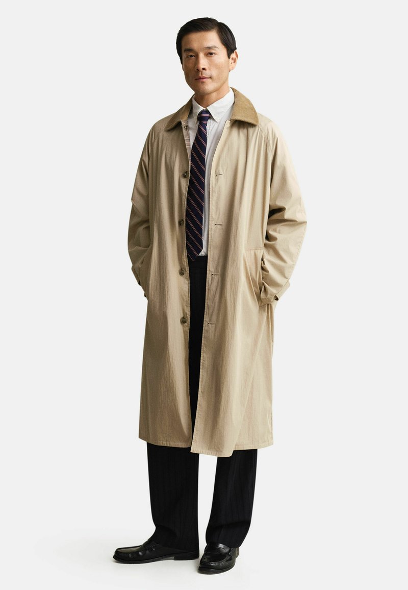 Mann trägt beigen langen Mantel mit braunem Kragen über weißem Hemd, gestreifter Krawatte, schwarzen Hosen und schwarzen Loafers, steht mit Händen in den Taschen.