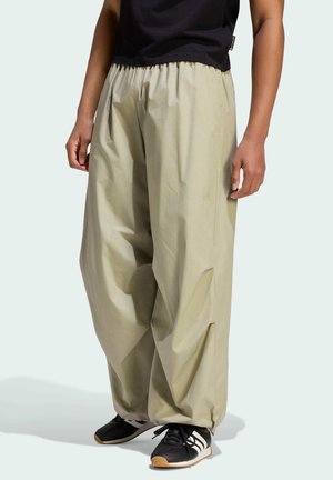 Persona vistiendo pantalones beige de pierna ancha con cintura y puños elásticos, zapatillas negras Adidas con rayas blancas y una camiseta negra.