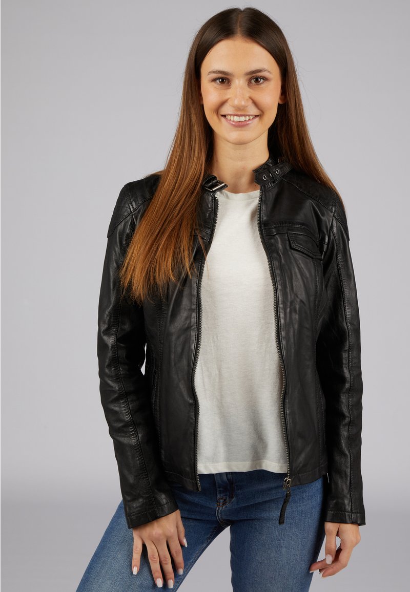 Schwarze Lederjacke mit tailliertem Design, mit Reißverschluss und dezenten Schulterakzenten, getragen über einem weißen Oberteil und blauen Jeans.