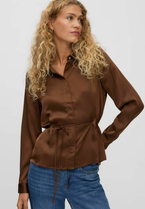 Femme aux cheveux blonds bouclés portant un chemisier marron à manches longues avec un lien à la taille et un jean bleu, regardant sur le côté.