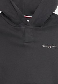 Hanorac cu glugă de culoare gri închis, dotat cu buzunar tip kangaroo. Prezintă un mic logo alb "TOMMY HILFIGER" și un detaliu în culorile roșu, alb și albastru la gât.