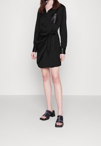 Robe chemise noire avec une taille nouée, manches longues et une poche poitrine en cuir. Associée à des sandales noires à talons blocs avec surpiqûres blanches.