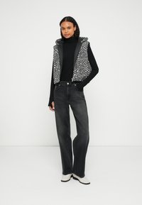 Αμάνικο γιλέκο με μαύρες και λευκές κηλίδες και κουκούλα πάνω από μαύρη μπλούζα με μακριά μανίκια, συνδυασμένο με ψηλόμεσα, φαρδιά τζιν παντελόνια σκούρου γκρι χρώματος και λευκά μποτάκια.