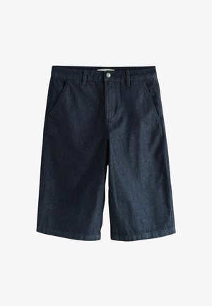 Femeie blondă cu păr ondulat, purtând top maro închis fără mâneci, pantaloni scurți închiși la culoare până la genunchi, ținând o geantă maro cu model, stând în fața unui perete gri.