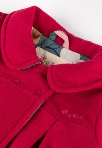 Cappotto in pile rosso con colletto tondo, caratterizzato da bottoni a pressione rosa e un marchio ricamato. L'interno presenta una fodera a fantasia in diverse colorazioni.
