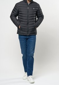 Svart puffjacka med horisontell sömnad, framdragkedja och två sidofickor. Bärs över en grå tröja och kombineras med blå jeans och vita sneakers.