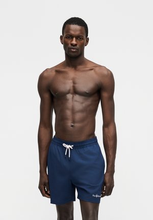 Μυώδης άντρας στέκεται προς τα εμπρός, φορώντας μπλε σκούρα μαγιό με λευκό κορδόνι και λογότυπο Polo Ralph Lauren στο αριστερό πόδι.