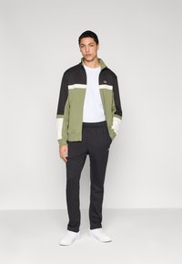 Veste de survêtement vert olive et noir avec des rayures blanches, assortie à un pantalon noir. Comprend une fermeture éclair et un détail logo. Baskets blanches.