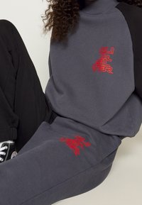 Persona con sudadera Quiksilver gris oscuro y pantalones a juego con el logo rojo, zapatillas negras, sentada con una rodilla levantada.