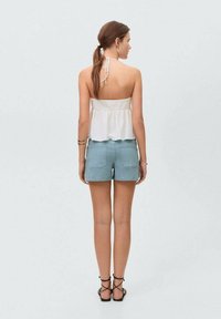 Bela halter majica s nabrano hrbtno stranico, v kombinaciji z svetlo modrimi jeans kratkimi hlačami. Opazne podrobnosti vključujejo zavezo okoli vratu in ravne sandale.