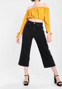 Gelbes, schulterfreies Crop-Top mit rüschendem Ausschnitt, kombiniert mit hoch taillierten schwarzen Flared-Jeans. Dazu werden schwarze High-Heels-Sandalen getragen.