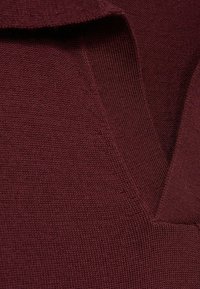 Materialul tricotat de culoare burgundă prezintă un design cu decolteu în V și o textură texturată. Marginile sunt finisate cu atenție, evidențiind textura sa moale.