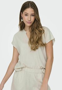 Jasno beżowa bluzka z krótkim rękawem, w dekolcie w kształcie litery V i elastycznym pasie. Wykonana z miękkiego, teksturowanego materiału. Prosty, luźny krój.