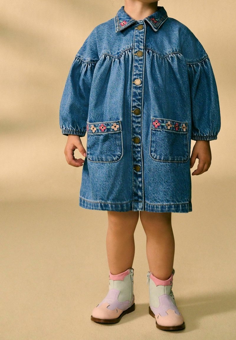 Enfant portant une robe en jean bleue avec une broderie florale sur le col et les poches, associée à des bottes de cowboy de couleur pastel, debout sur un sol beige.