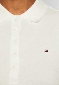 Valkoinen poolopaita, jossa on teksturoitu kangas, kolmonen napitus ja pieni brodeerattu logo punaisella, valkoisella ja sinisellä rinnassa.