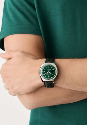 Homme en chemise verte, les bras croisés, portant une montre-bracelet argentée avec cadran vert et bracelet en cuir noir au poignet gauche.