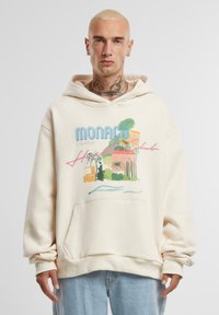 Béžová mikina s kapucí, která má grafický potisk tropické scény se textem "Monaco" a "Côte d'Azur", texturovanou látku a přední kapsu.