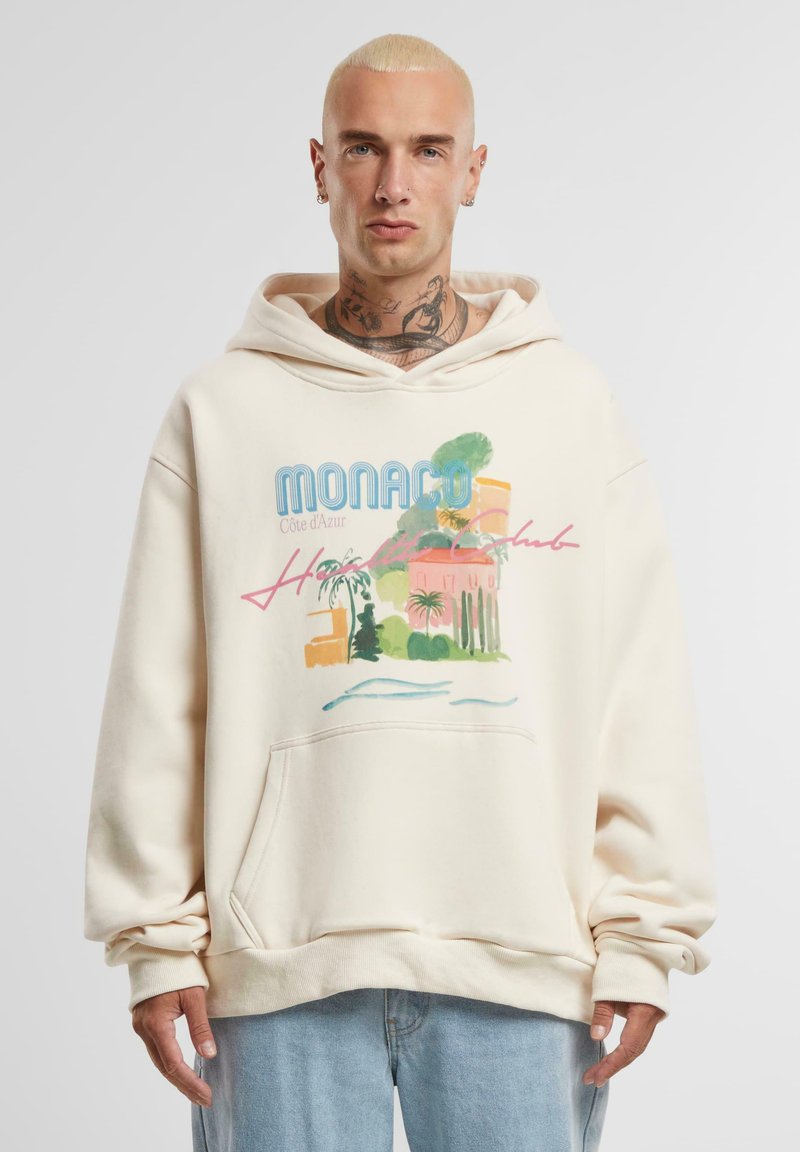 Béžová mikina s kapucí, která má grafický potisk tropické scény se textem "Monaco" a "Côte d'Azur", texturovanou látku a přední kapsu.