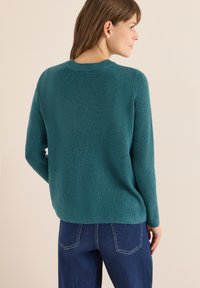 Turkusowy sweter dziany o prążkowanej fakturze, z okrągłym dekoltem i długimi rękawami, w zestawieniu z niebieskimi dżinsami z kontrastowymi szwem.