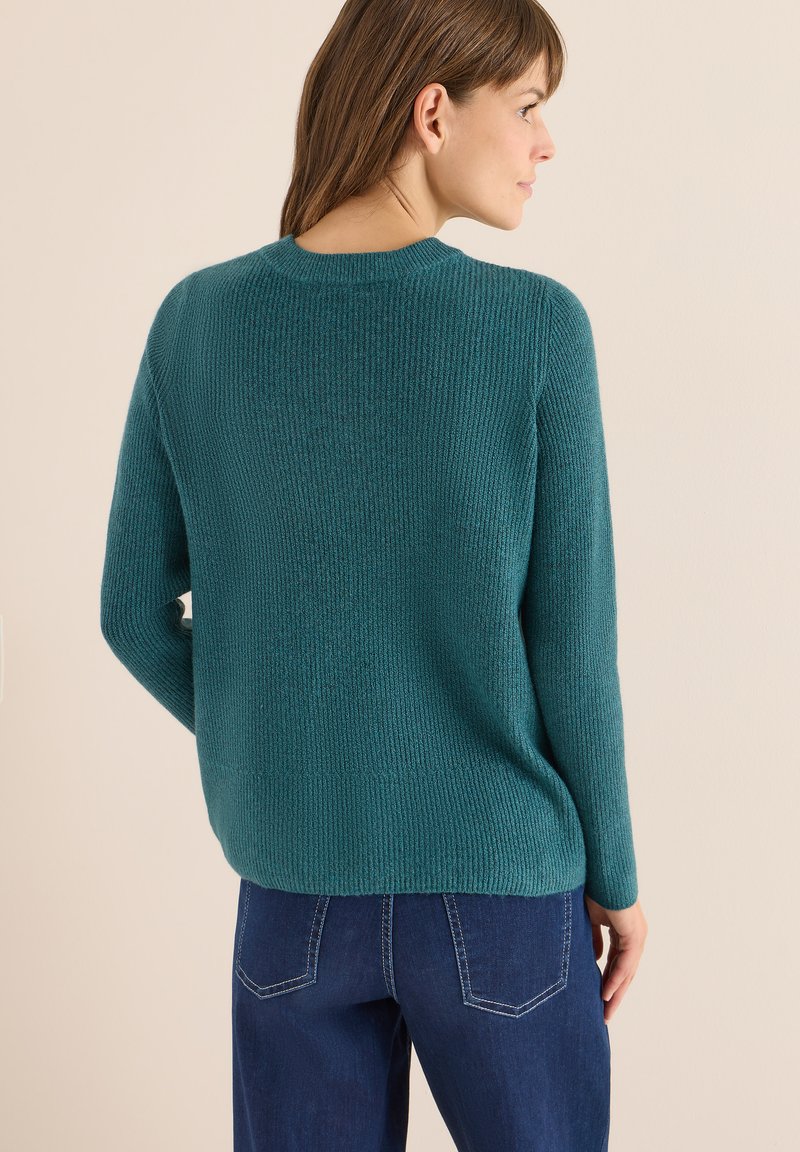 Pull en tricot teal avec une texture côtelée, un col rond et des manches longues, associé à un jean en denim bleu avec des coutures contrastées.