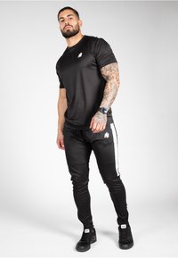 Black athletic shirt and pants, both made of smooth fabric. Shirt features a small white logo; pants have white side stripes and a logo.