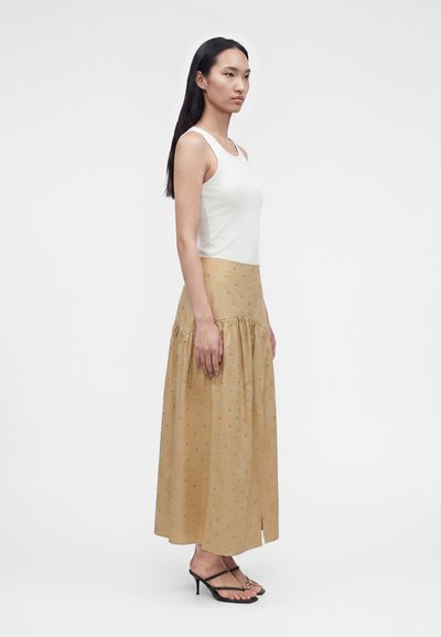 Γυναίκα που στέκεται πλαγίως, φοράει άσπρο αμάνικο τοπ, μακριά μπεζ φούστα με σχέδια και μαύρα σανδάλια με τακούνι σε μονόχρωμο φόντο.