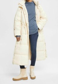 Puffjacka i ljusbeige med hög krage, matchad med en blå tröja och mörka jeans. Beiga ankelstövlar med texturerade sulor.