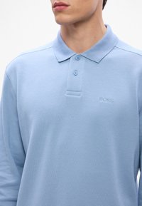 Gaiši zilas polo krekls ar strukturētu audumu, kuram ir apkakle, trīs pogu aizdare un izšūts "BOSS" logo uz krūšu daļas.