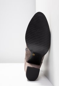 Semelle d'une botte en daim marron avec semelle extérieure et talon en caoutchouc noir texturé, placée debout contre un mur d'angle blanc. Taille 37 marquée sur la semelle.