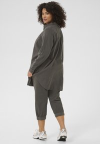Chemise oversize vert olive à manches longues et poche poitrine, assortie à un pantalon court. Portée avec des chaussures de sport grises.