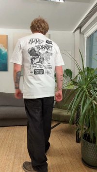 Vit bomullst-shirt med grafiskt tryck på baksidan, svart bläckillustrationer och text. Kombinerad med svarta byxor och bruna skor.
