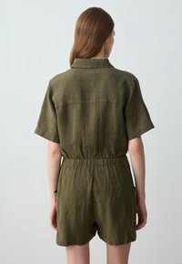 Olivgrüner Leinen-Jumpsuit mit kurzen Ärmeln, elastischem Bund und seitlichen Taschen. Mit Kragen und lässiger Passform. Rückansicht gezeigt.