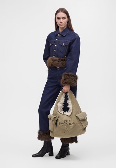 Femme en veste et pantalon en jean bleu foncé avec bordure en fourrure marron tenant un grand sac fourre-tout beige étiqueté "The Sack Bag Marc Jacobs."