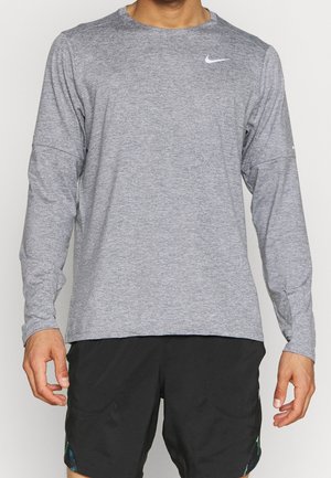 Homme portant un t-shirt Nike gris à manches longues et un short noir, debout devant un fond clair uni.