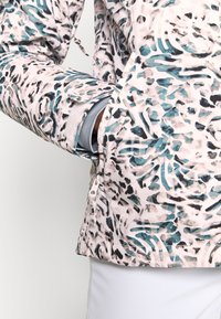 Personne portant une veste rose clair à motifs avec des dessins abstraits turquoise et noirs, mains partiellement dans les poches de la veste.